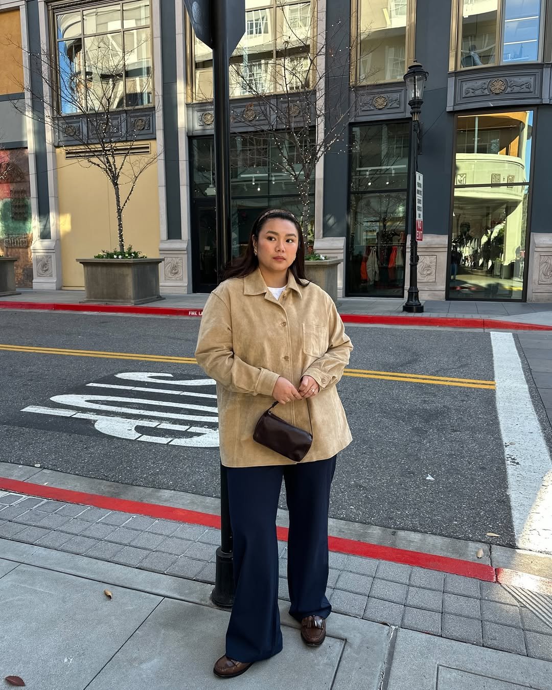 Pessoa de pé na calçada, vestindo casaco bege e segurando bolsa. Ela está em uma rua urbana com prédios ao fundo.