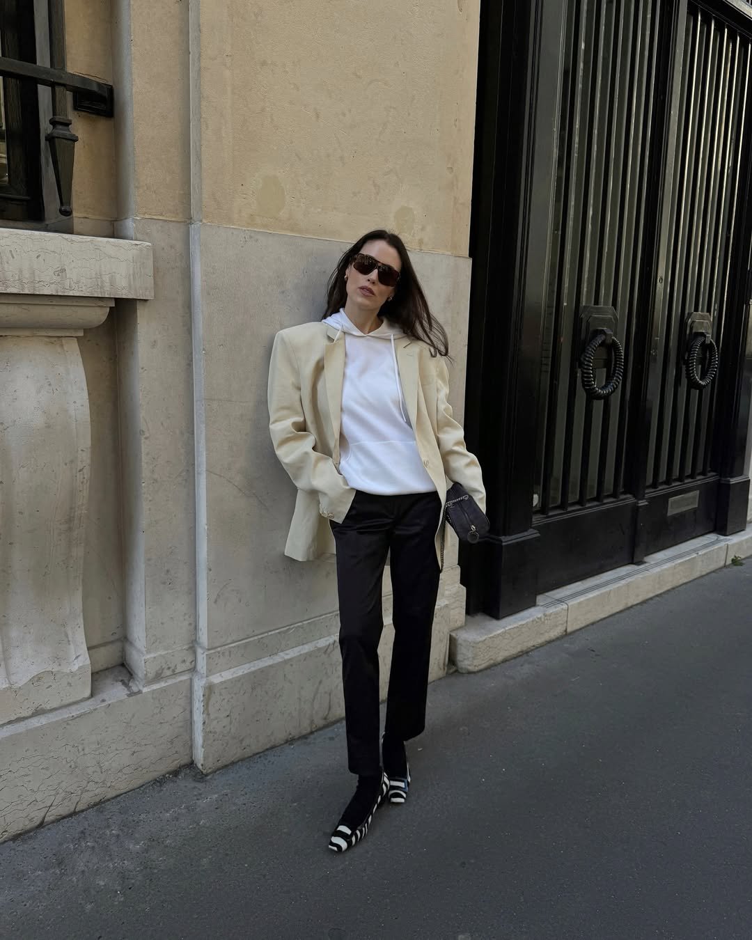 Pessoa de óculos escuros encostada em prédio, usando blazer claro e calça escura, estilo moderno para entrevista de emprego.