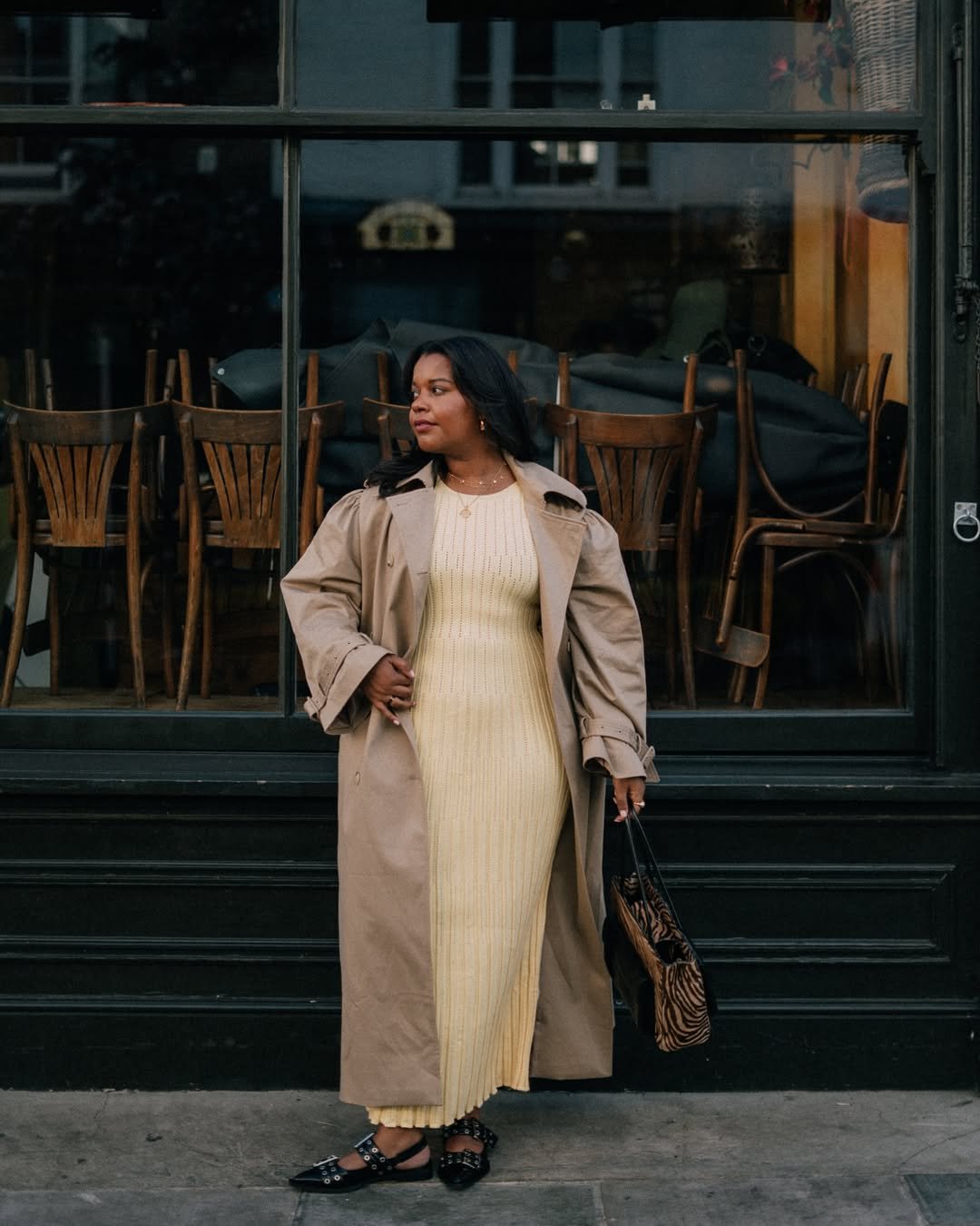 Mulher em frente a uma vitrine usando um vestido longo amarelo e casaco bege, segurando uma bolsa listrada.