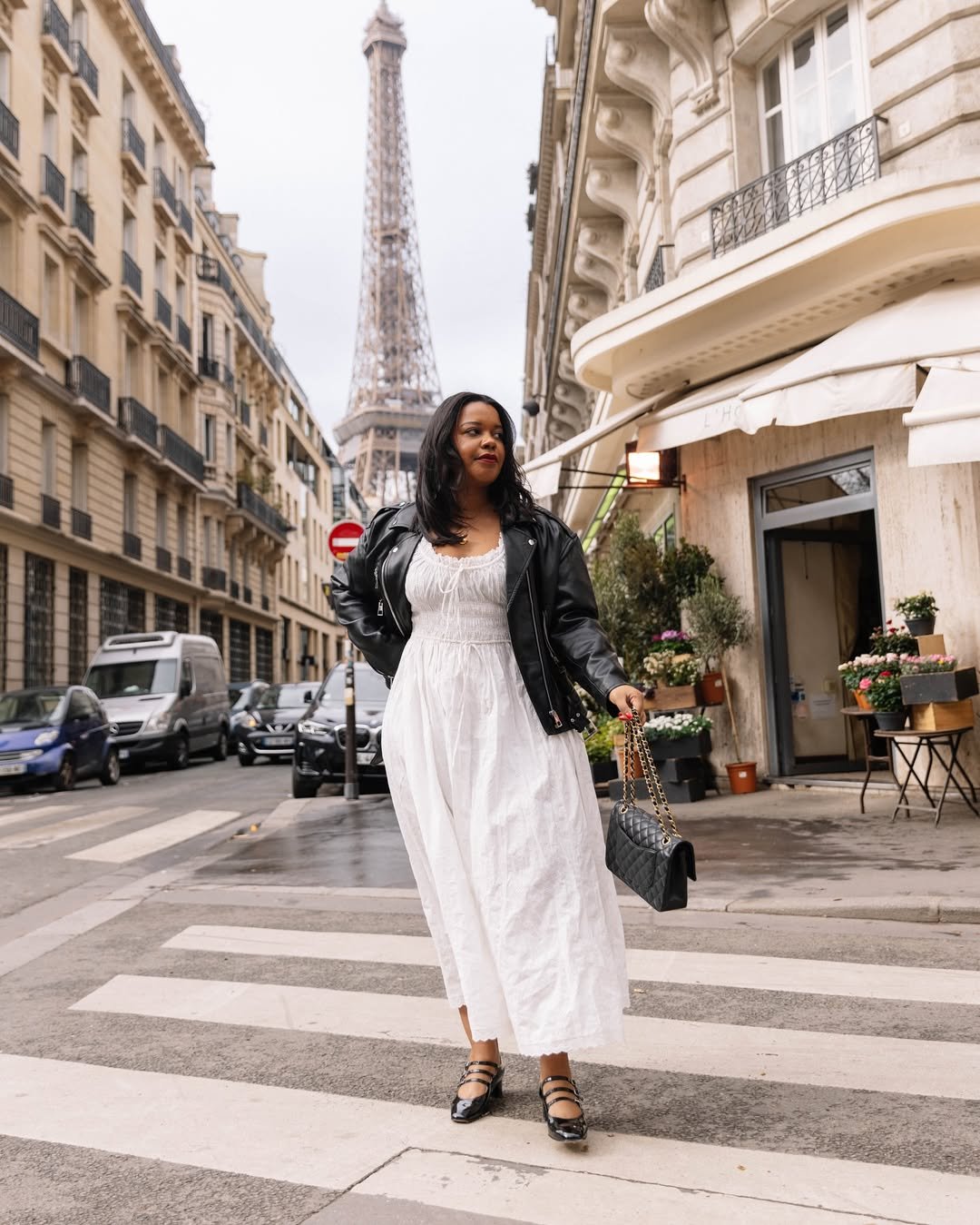 Mulher com vestido longo branco e jaqueta preta cruza a rua com a Torre Eiffel ao fundo.