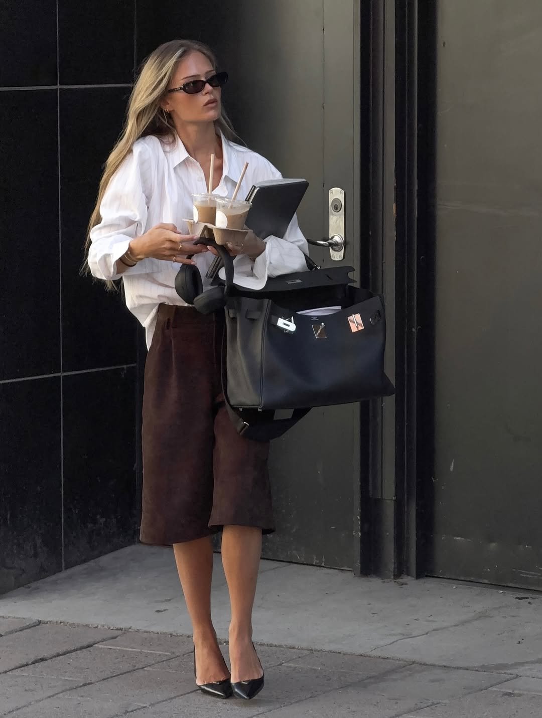 Pessoa elegante carrega um laptop e bebidas em direção a uma porta, possivelmente após uma entrevista de emprego.