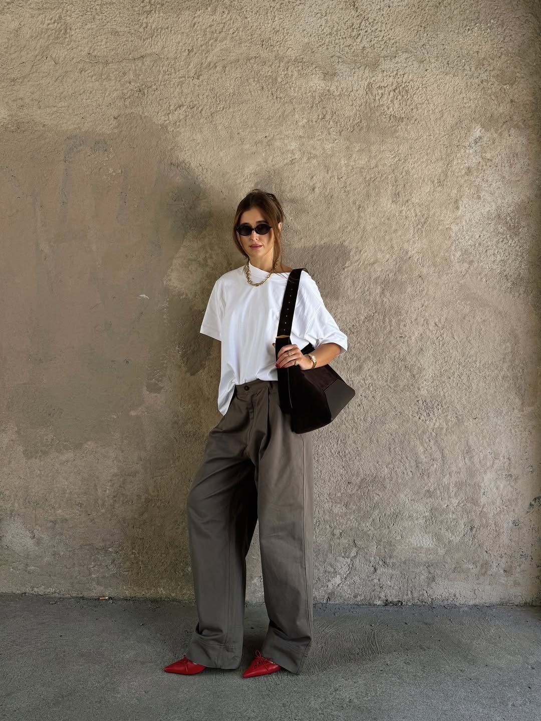 Pessoa em pé vestindo camiseta branca e calça larga, com bolsa e óculos escuros.