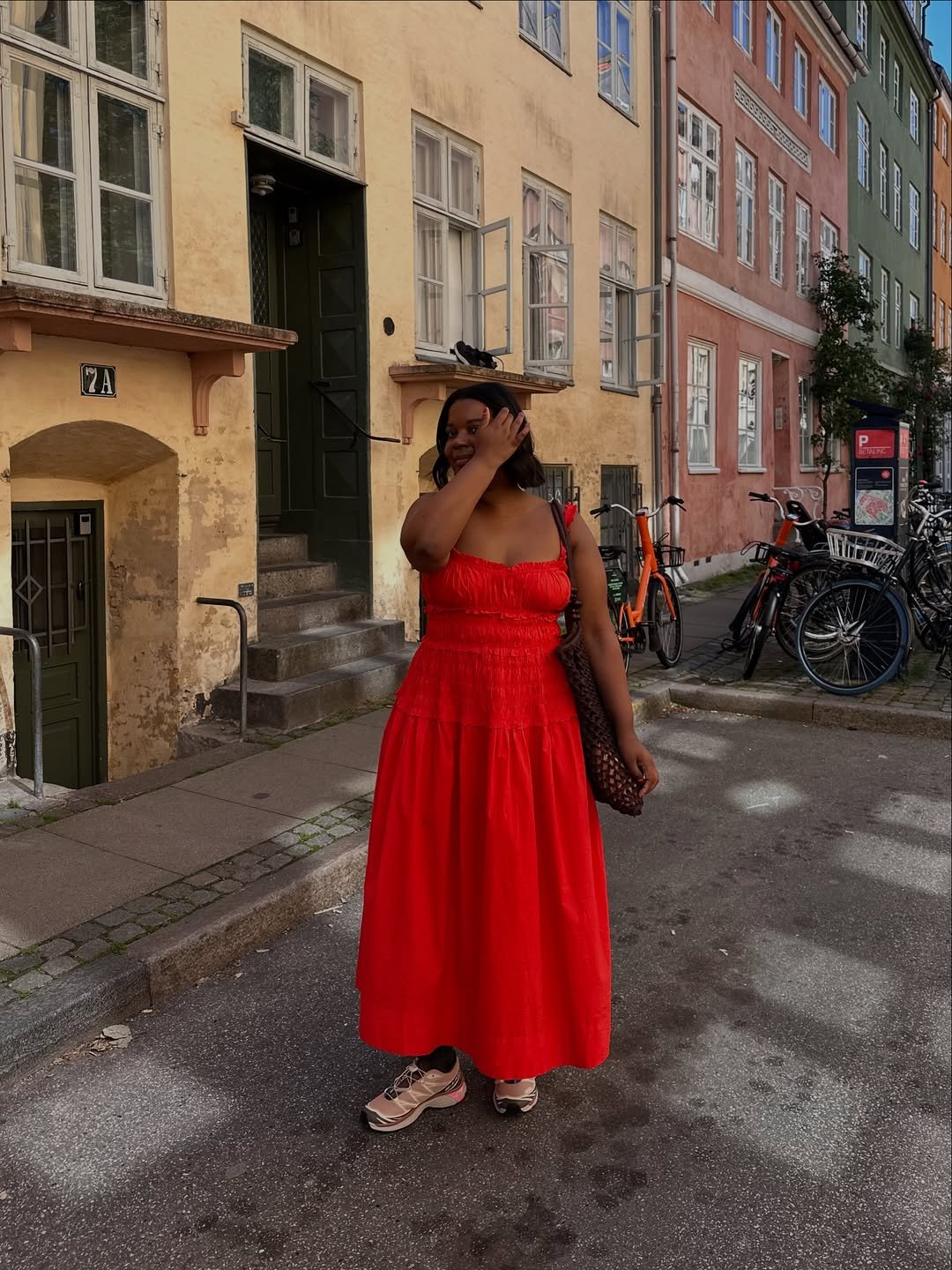 Pessoa usando um vestido longo vermelho em uma rua com bicicletas estacionadas e edifícios coloridos ao fundo.