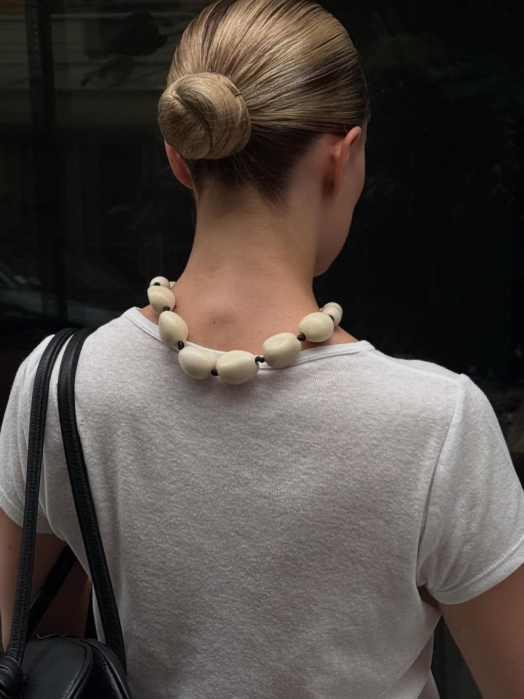 Uma mulher de coque elegante e colar de pedras brancas, vestindo camiseta branca, segura uma bolsa preta. Estilo formal para entrevista de emprego.