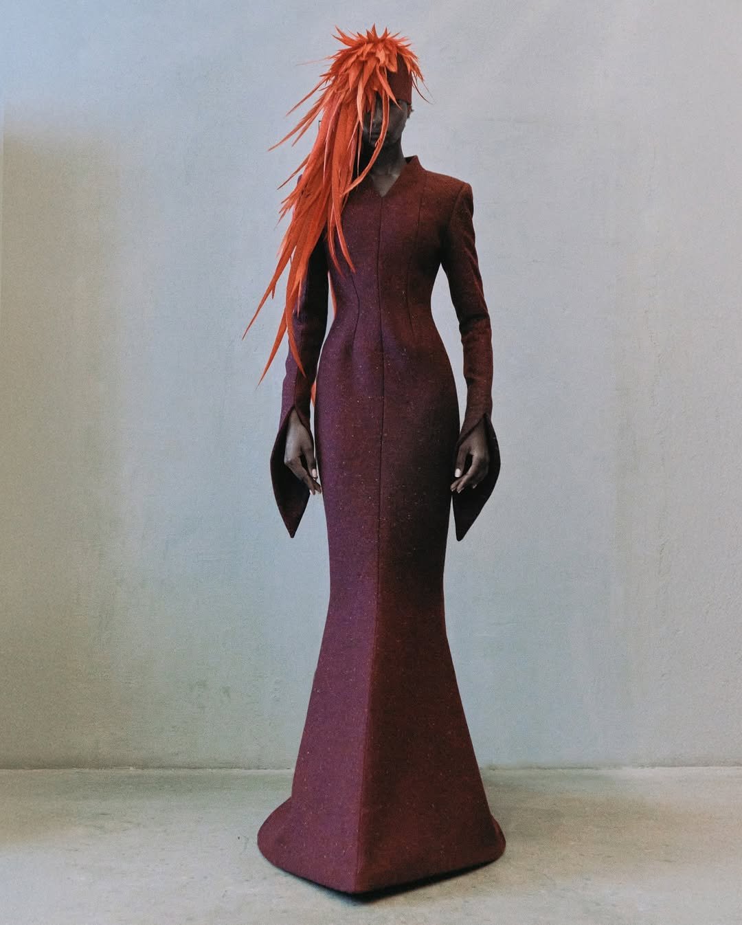 Vestido vinho elegante com mangas amplas e adorno capilar vermelho vibrante, em um cenário simples, evocando Schiaparelli.