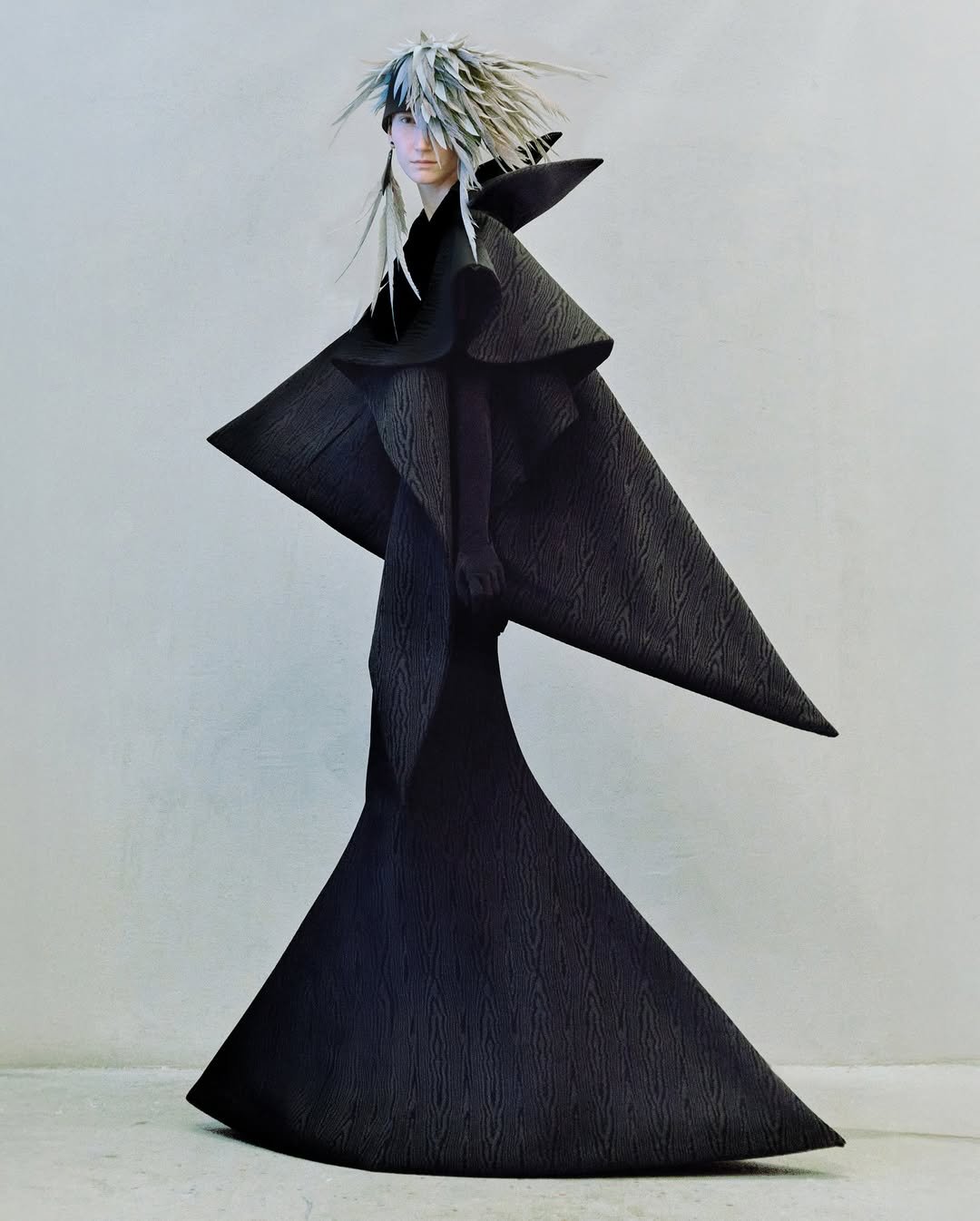 Pessoa em traje avant-garde preto com textura distinta e cabelo espetado prateado, estilizado de forma futurista.