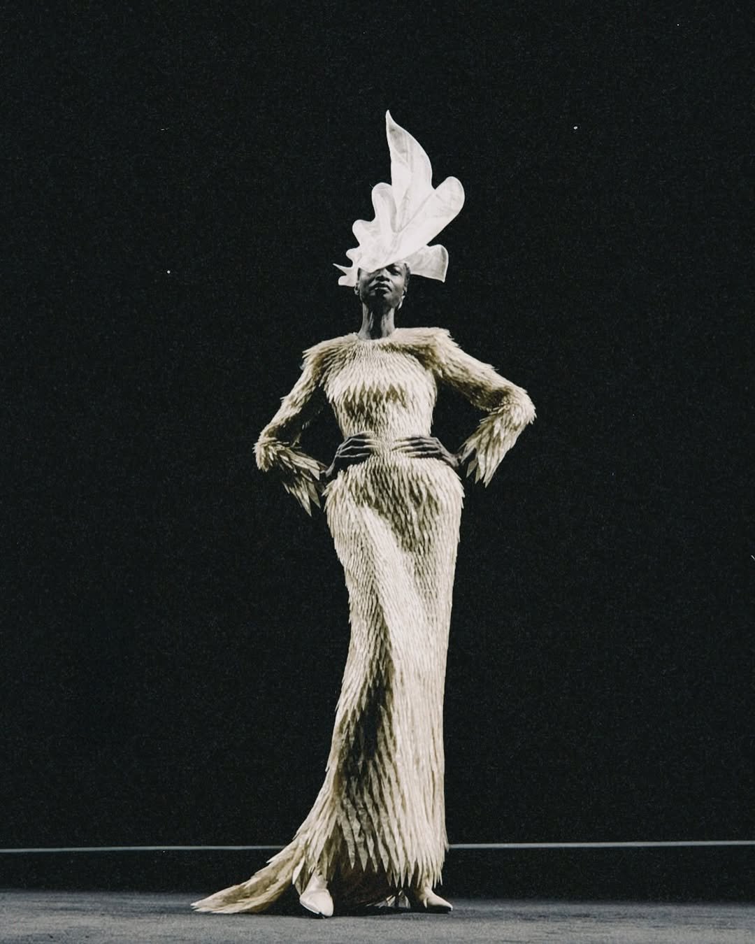 Vestido texturizado com estilo avant-garde e adorno de cabeça escultural em destaque em fundo escuro.