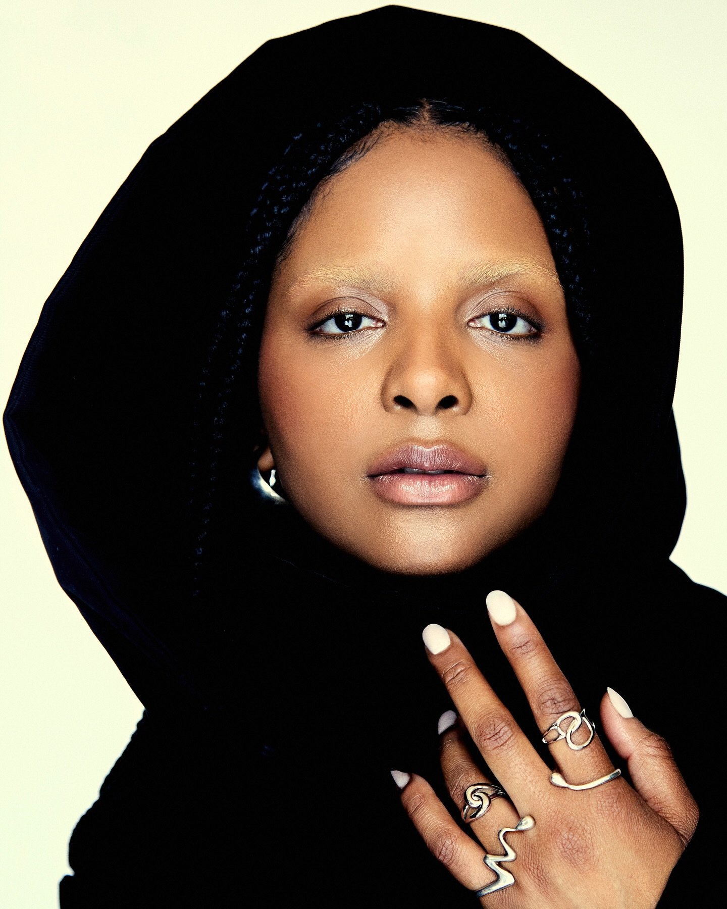 Pessoa com capuz preto e sobrancelhas descoloridas, usando anéis elegantes nas mãos levantadas.