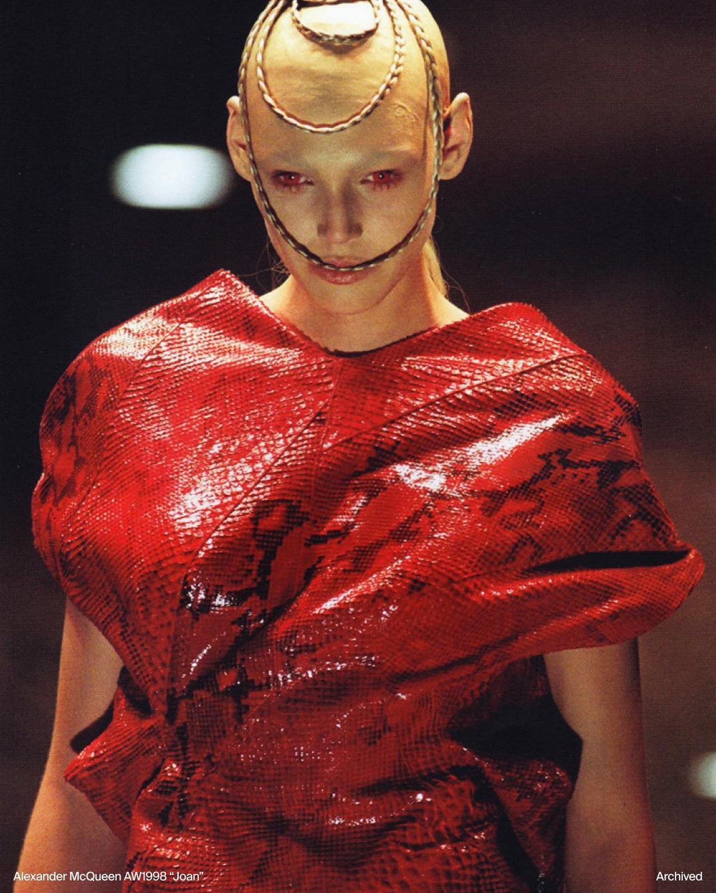 Modelo com sobrancelhas descoloridas veste roupa vermelha texturizada; adorno de cabeça metálico.