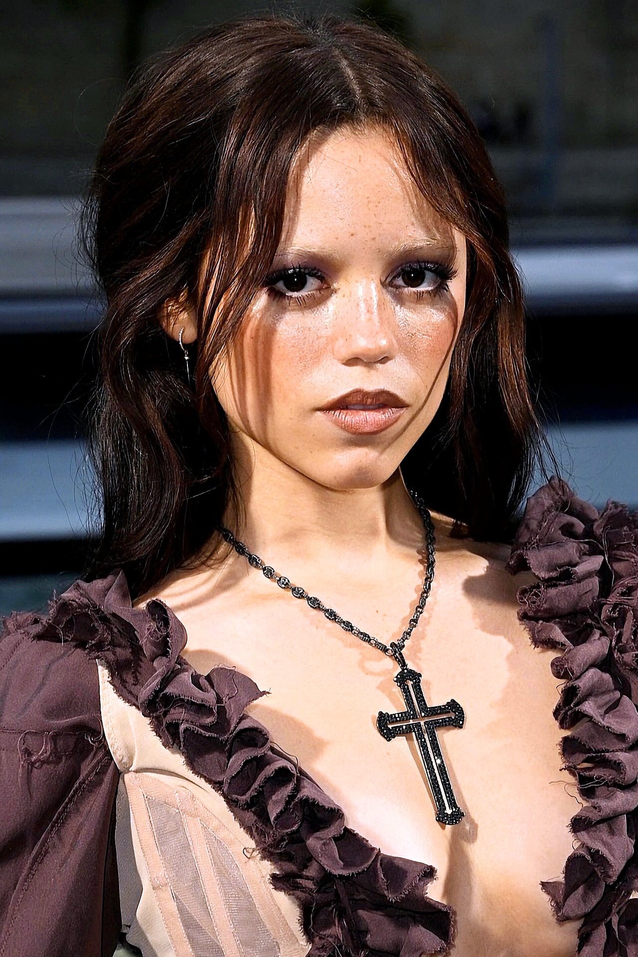 Pessoa com sobrancelhas descoloridas, vestindo colar com pingente de cruz e roupa de babados marrons.