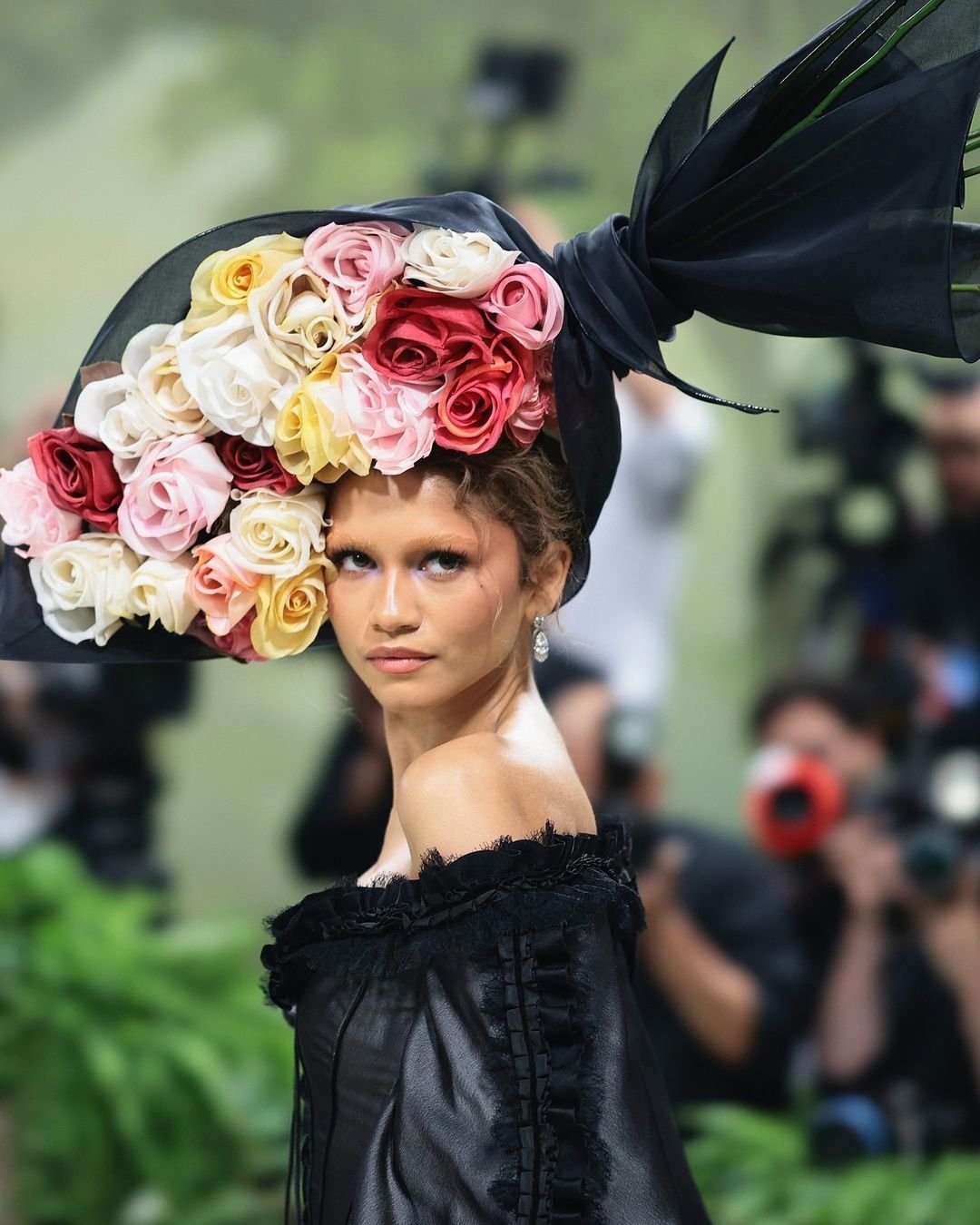 Modelo com sobrancelha descolorida, usa vestido preto e chapéu espetacular com flores coloridas.
