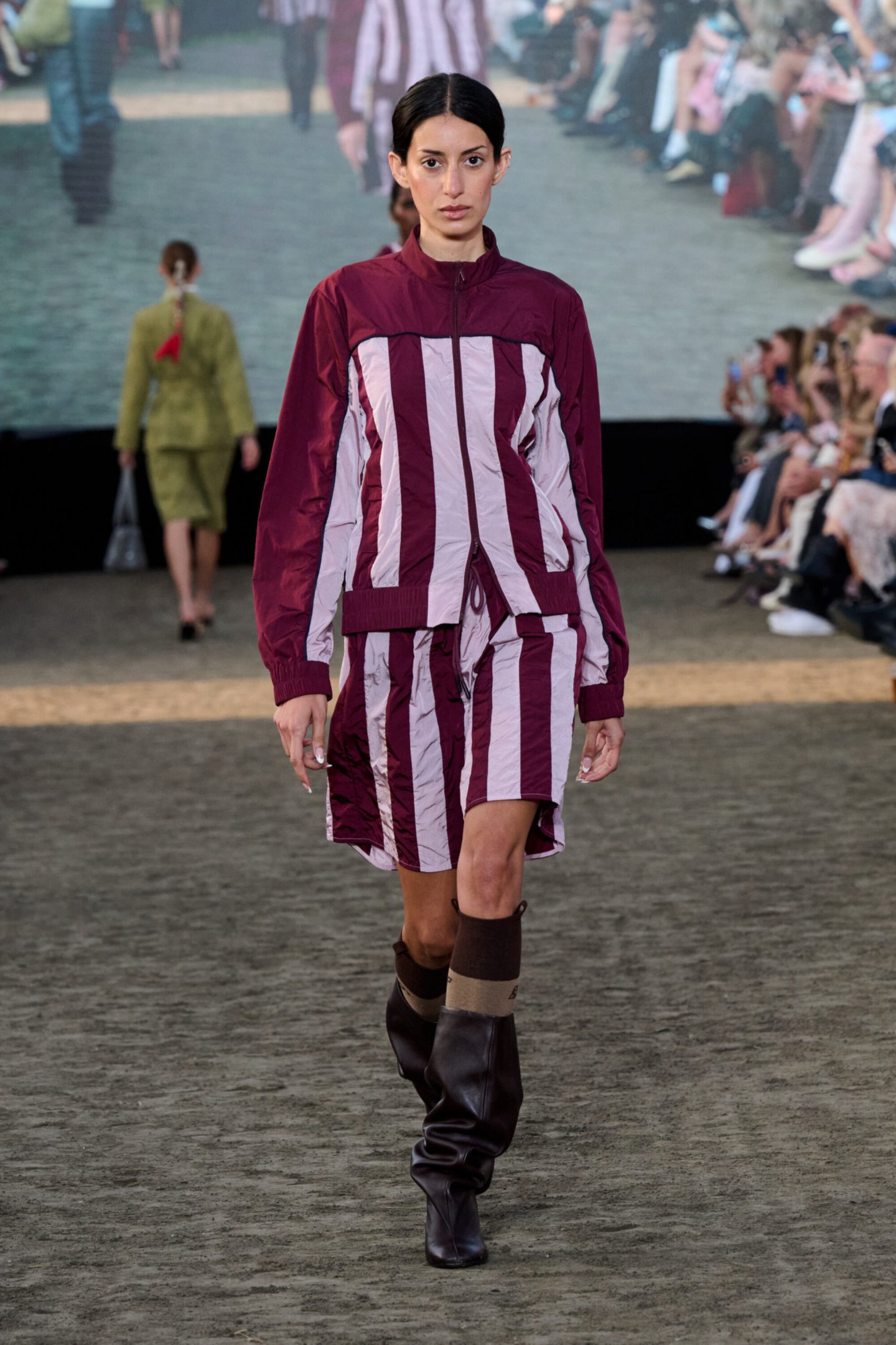 Desfile da Copenhagen Fashion Week Spring 2026 com modelo usando conjunto listrado bordô e botas pretas.