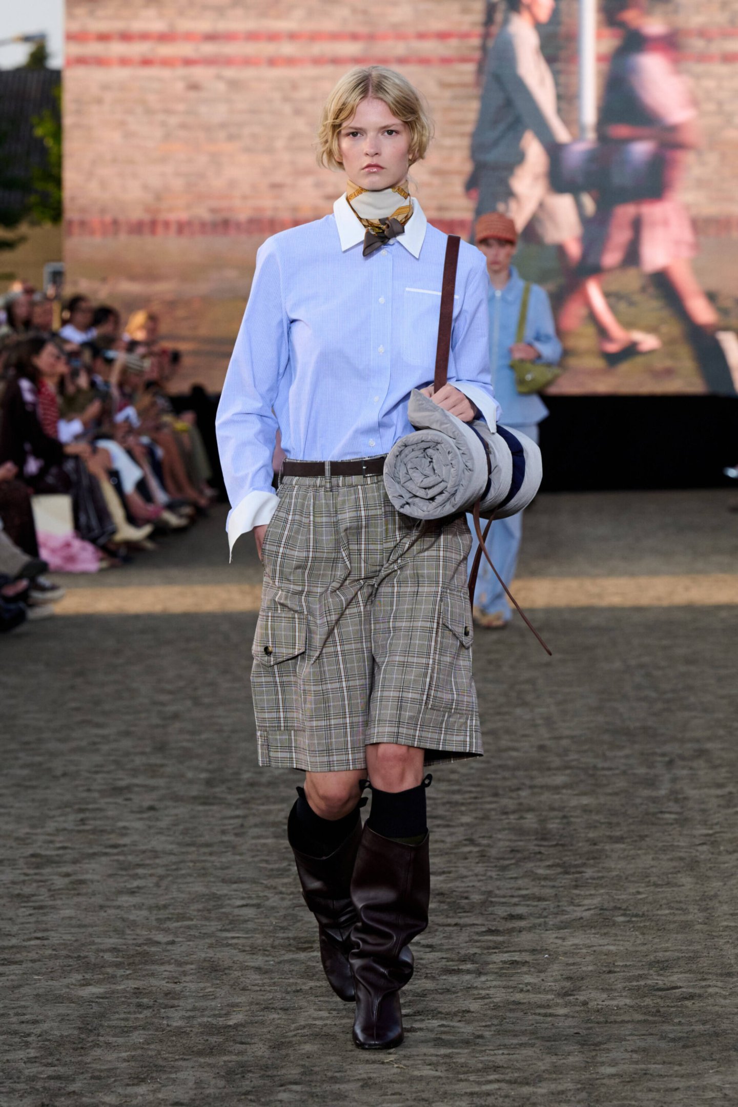 Modelo em desfile com camisa azul clara, bermuda xadrez e botas de couro escuras durante a Copenhagen Fashion Week Spring 2026.