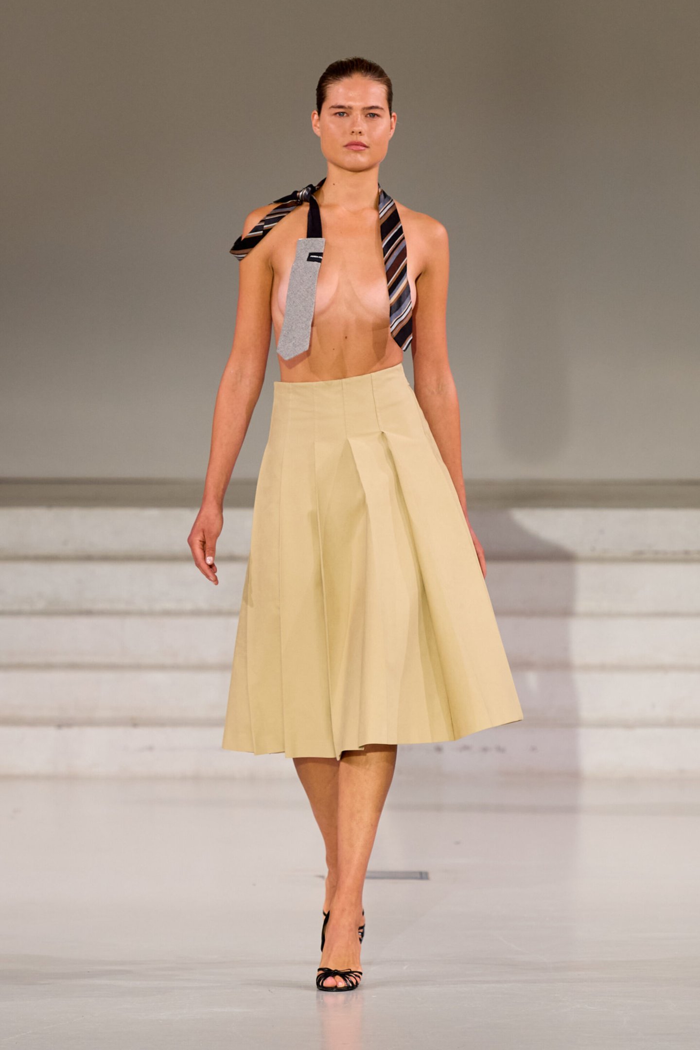 Modelo desfilando na Copenhagen Fashion Week Spring 2026 com saia bege e acessório de gravata.