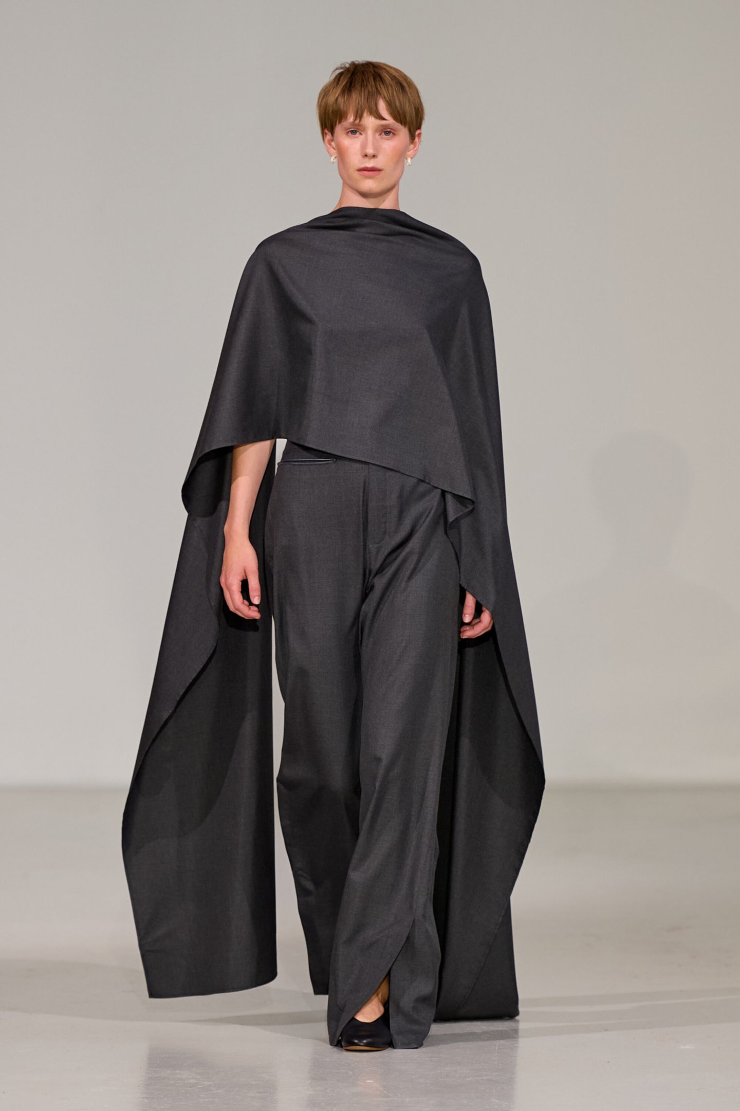 Modelo em passarela com conjunto cinza e capa ampla na Copenhagen Fashion Week Spring 2026.