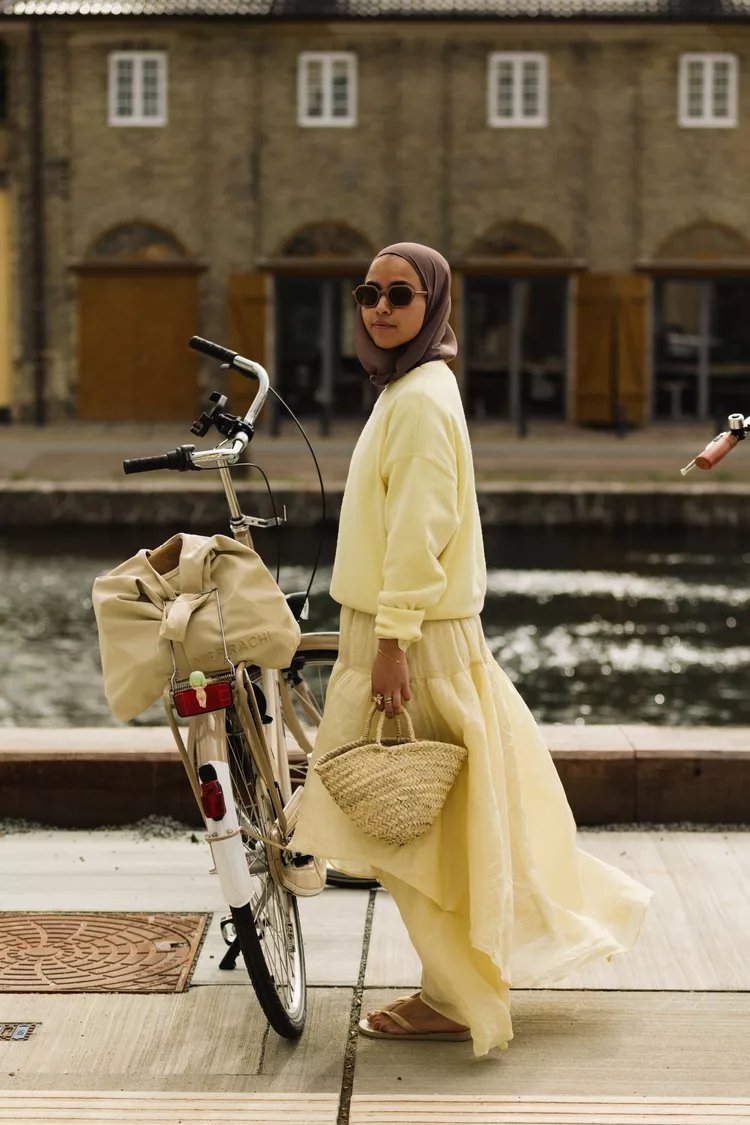 Pessoa com hijab e óculos escuros ao lado de bicicleta, usando roupa amarela e bolsa de palha. Estilo urbano na Copenhagen Fashion Week.