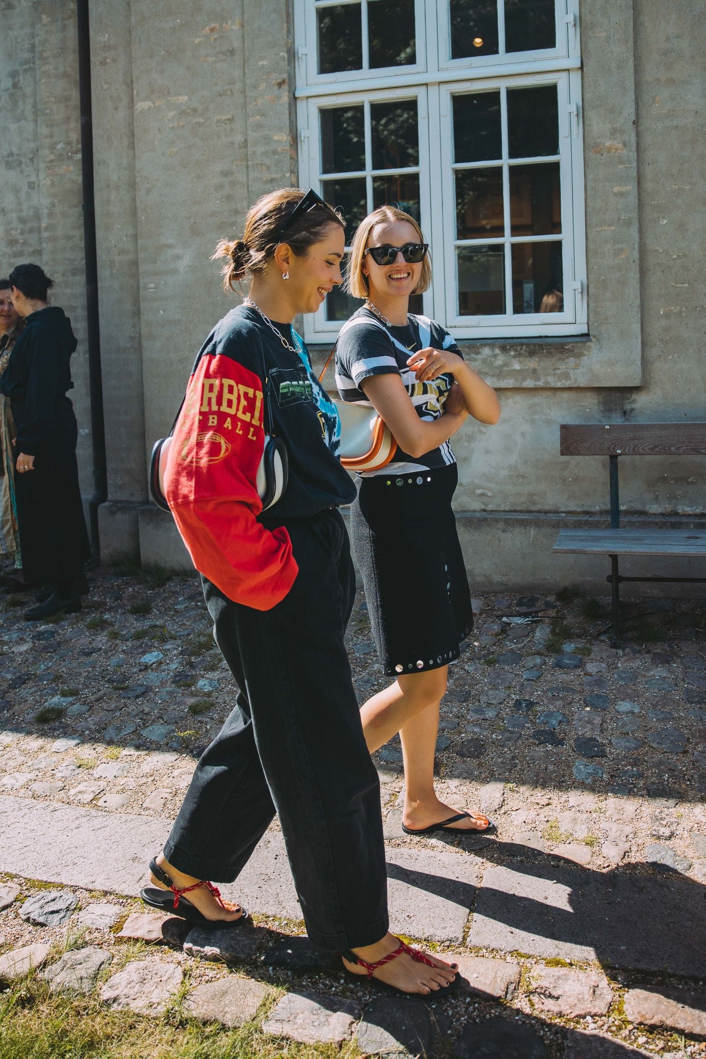 Duas mulheres sorriem e caminham, vestindo roupas casuais e estilosas durante a Copenhagen Fashion Week.