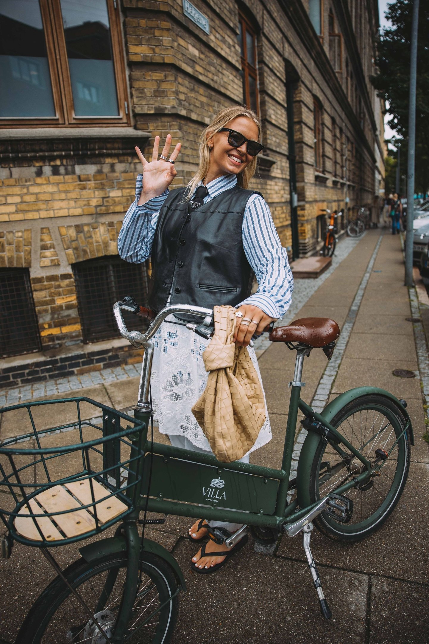 Mulher sorrindo em bicicleta, vestindo camisa listrada e colete preto, exemplificando street style da Copenhagen Fashion Week.