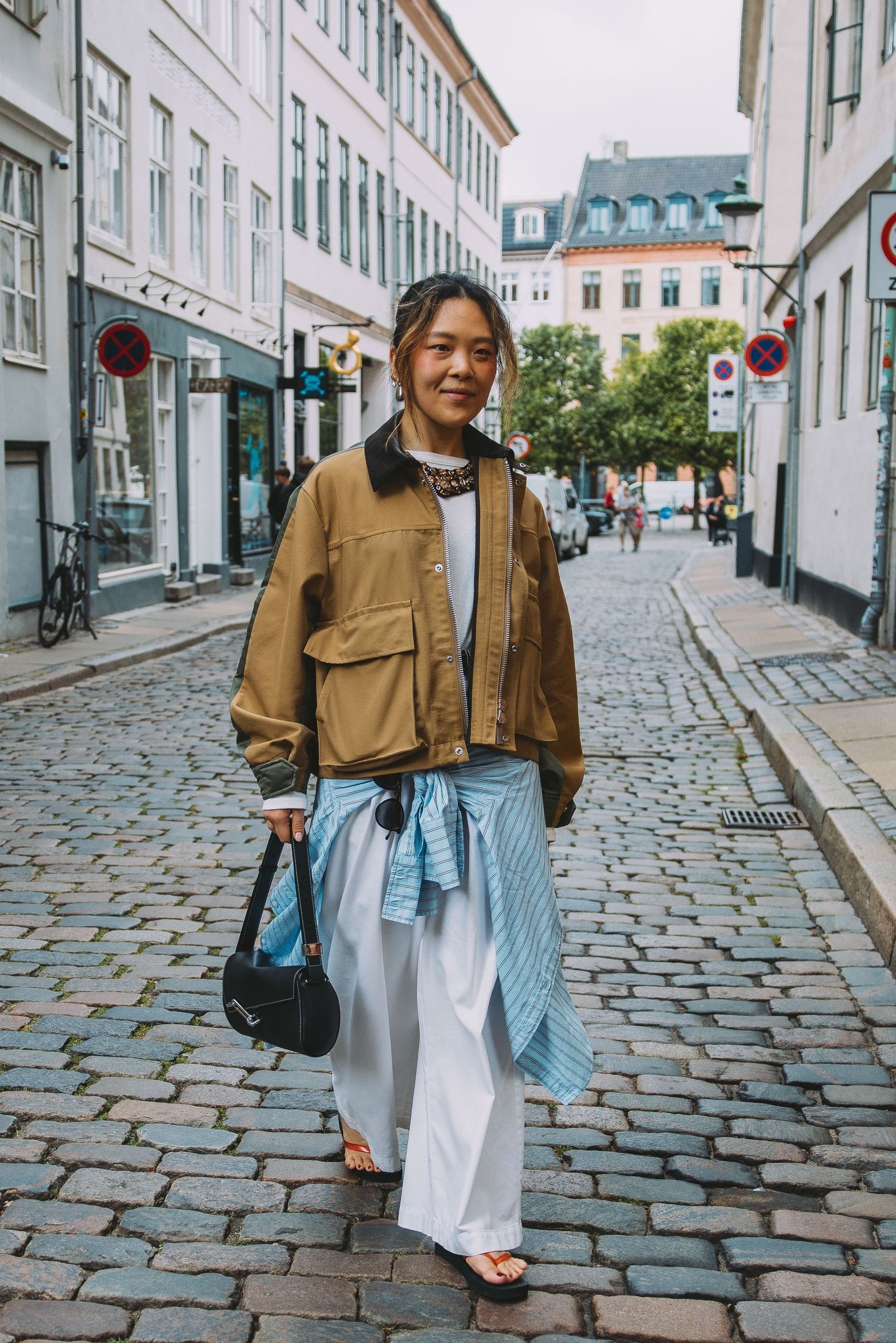 Mulher com estilo urbano em rua de paralelepípedos na Copenhagen Fashion Week, usando jaqueta cáqui e calças largas.