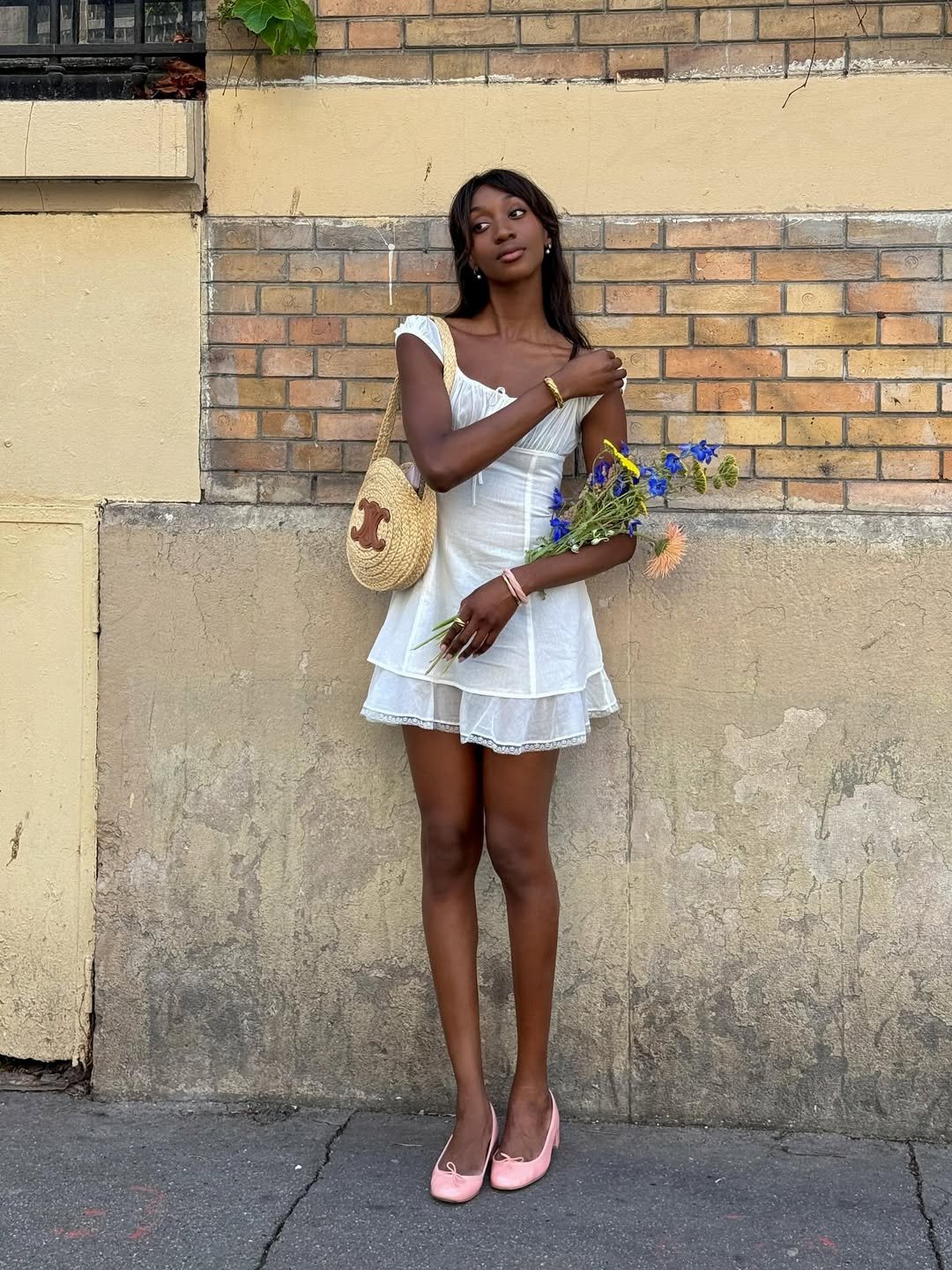 Jovem em vestido branco, segurando flores, com bolsa de palha, refletindo tendências verão 2026.