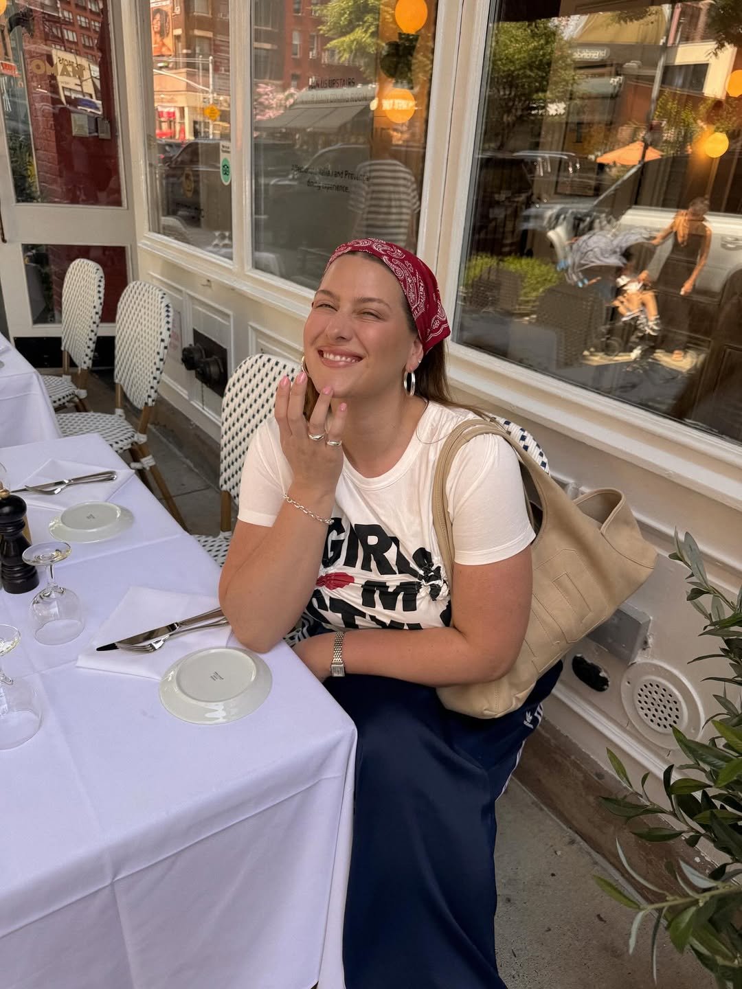Pessoa sorridente com lenço vermelho na cabeça, sentada em restaurante ao ar livre. Tendências verão 2026.