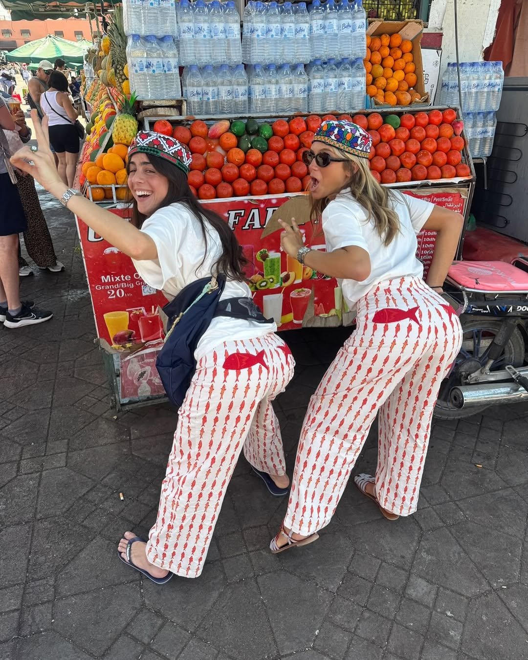 Duas pessoas sorridentes com calças estampadas em frente a uma banca de frutas, sinal de tendências verão 2026.