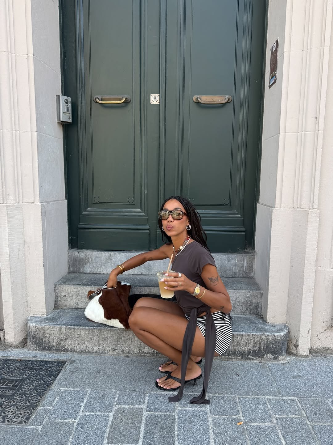 Pessoa com óculos de sol, blusa marrom e sandálias, sentada na porta, exemplificando tendências verão 2026.