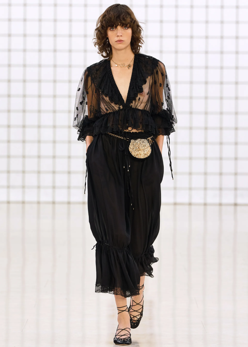 Modelo usa blusa preta transparente e calça bloomer, detalhes com renda e bolsa dourada na passarela.