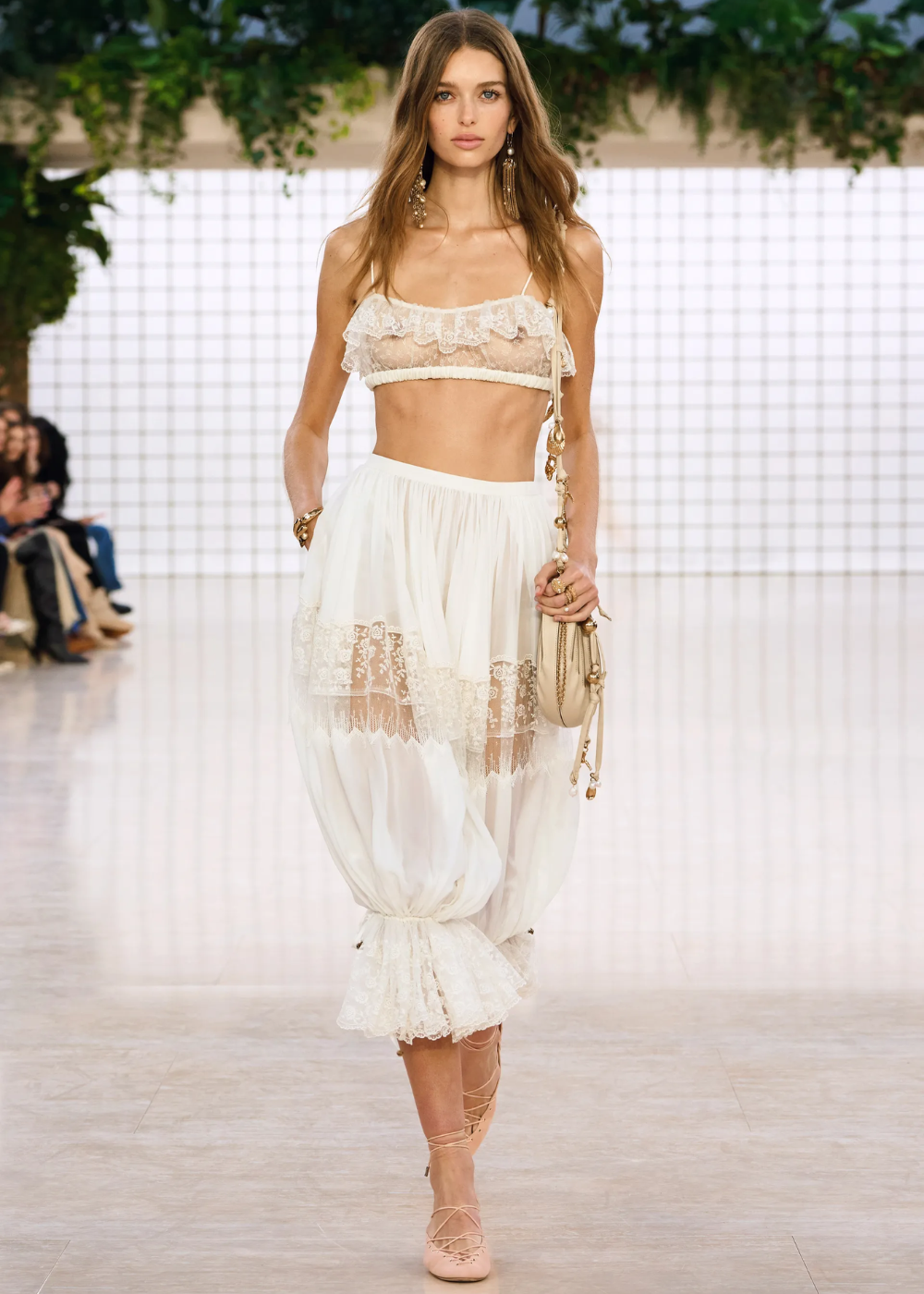 Modelo desfilando com conjunto leve de renda, incluindo top e calça estilo bloomer, em passarela iluminada.