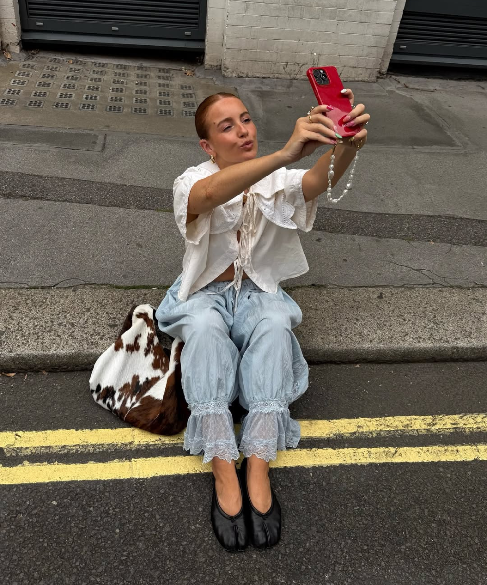 Pessoa sentada na calçada, vestindo calça estilo bloomer azul claro e blusa branca, tirando selfie com celular vermelho.