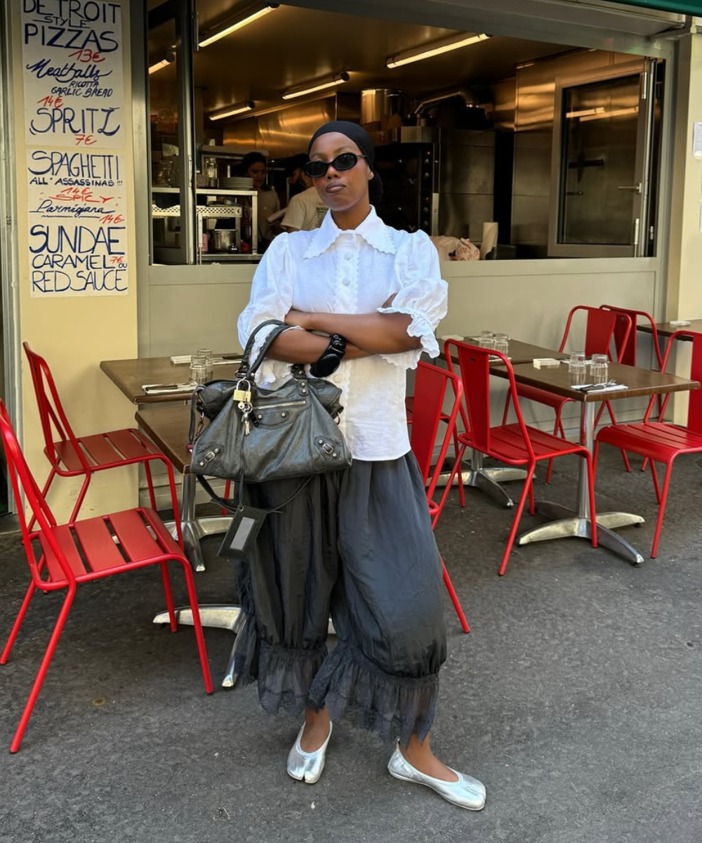 Pessoa de pé usando blusa branca, calça bloomer preta e sapatos prateados; segurando bolsa em frente a um café com cadeiras vermelhas.