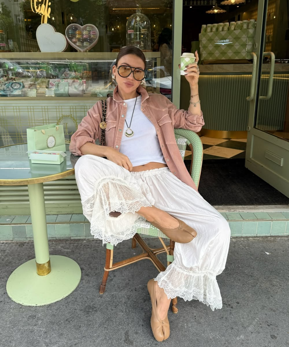 Pessoa usando calça bloomer branca com renda, casaco rosa e tomando café em frente a uma confeitaria.
