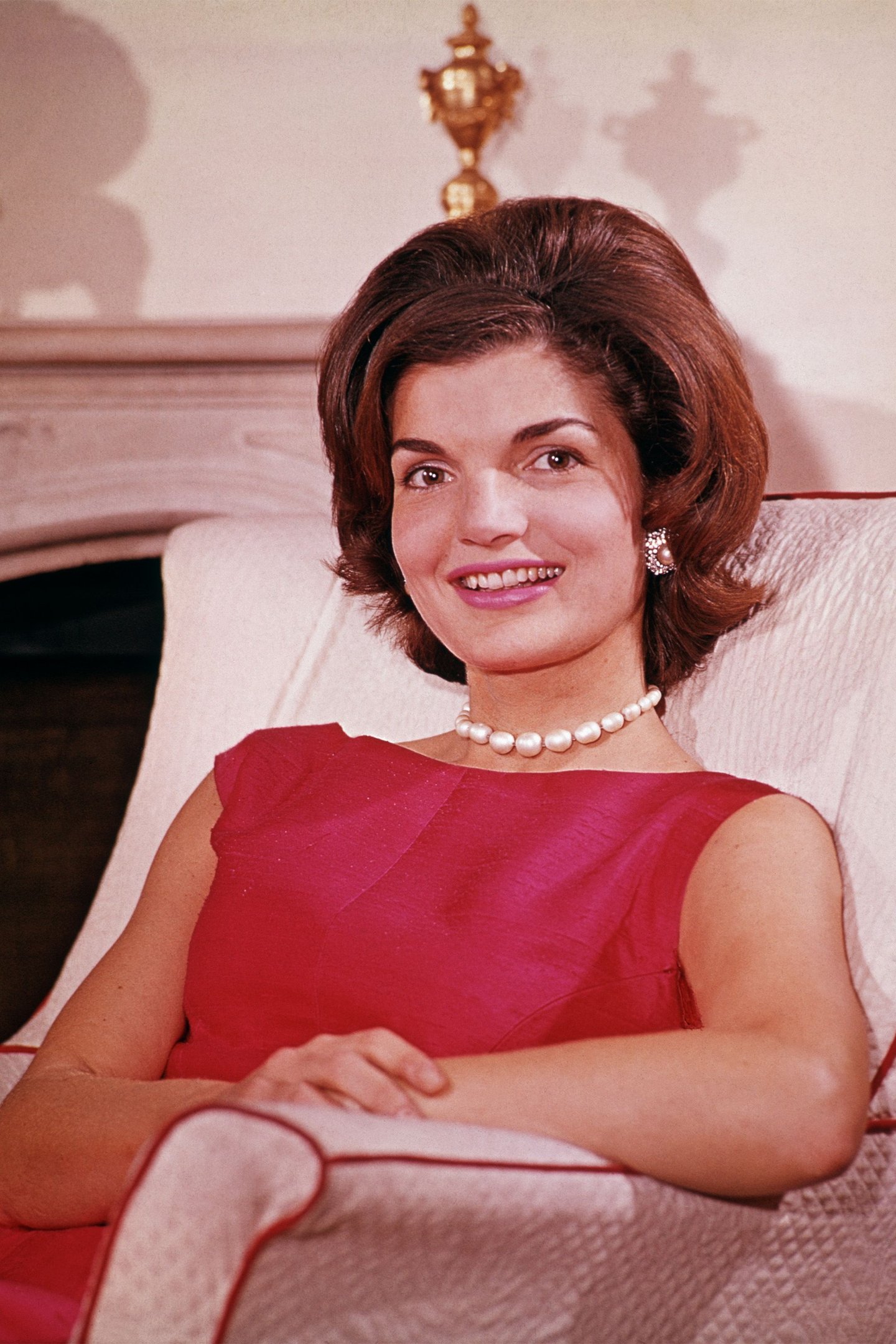 Mulher com vestido vermelho e colar de pérolas sentada em uma poltrona elegante.