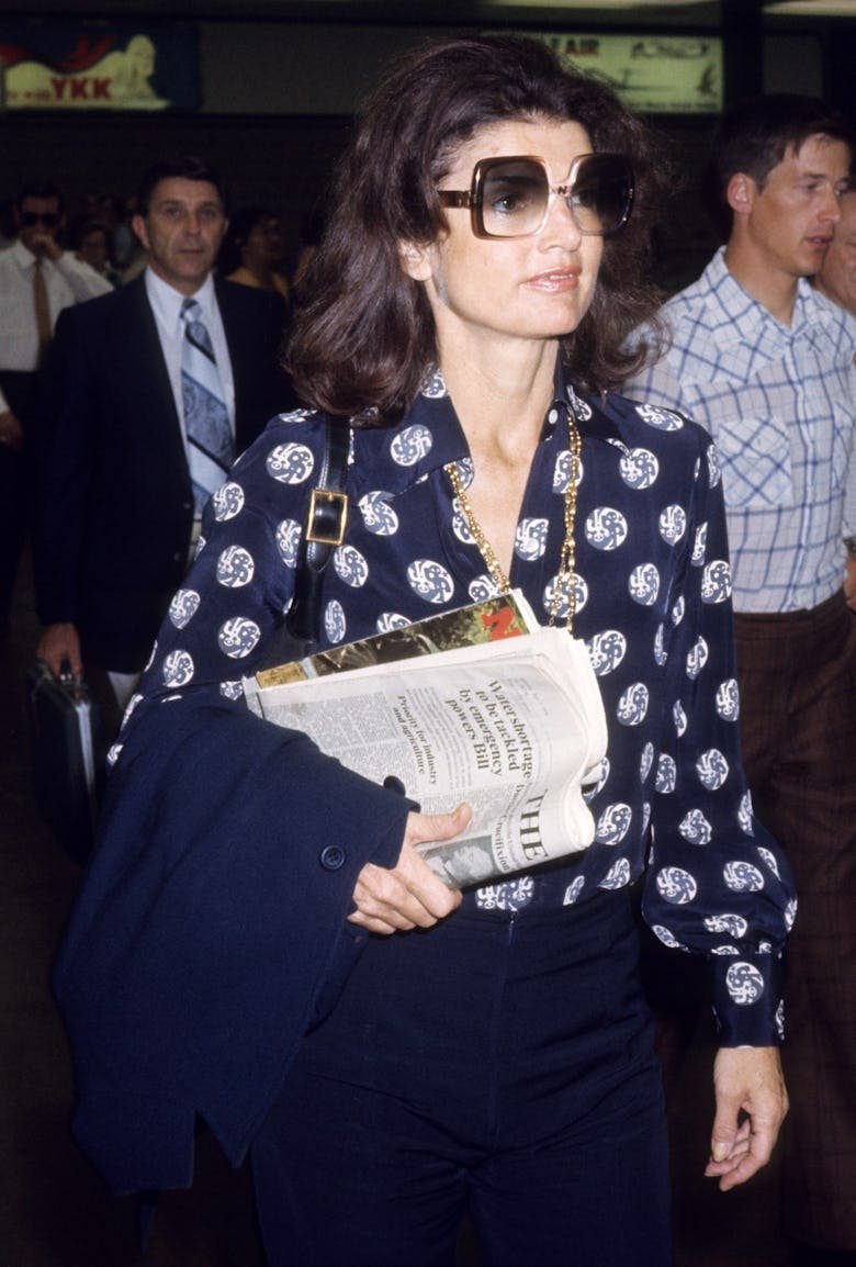 Mulher com óculos grandes vestindo camisa estampada, segura jornal e casaco, em ambiente movimentado.