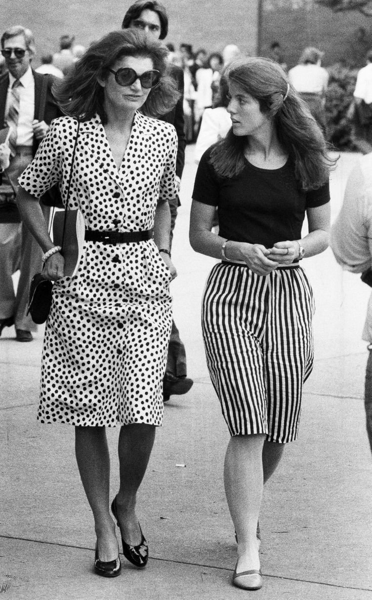 Duas mulheres caminhando lado a lado em meio a uma multidão. A da esquerda usa vestido de bolinhas e óculos escuros.