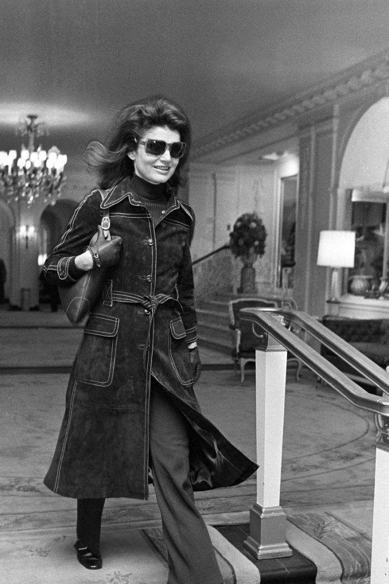 Mulher de casaco escuro e óculos escuros, subindo uma escada em um ambiente elegante.