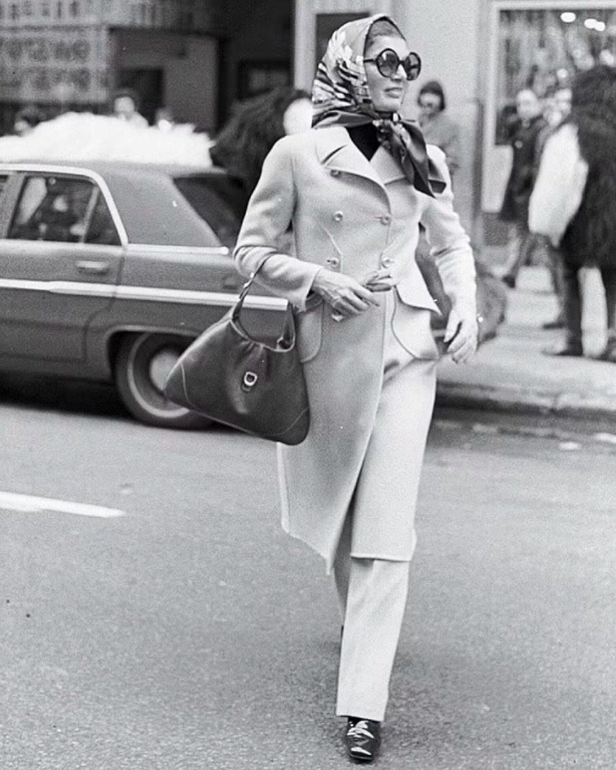 A imagem mostra uma mulher elegante vestindo sobretudo, óculos escuros e lenço, carregando bolsa.