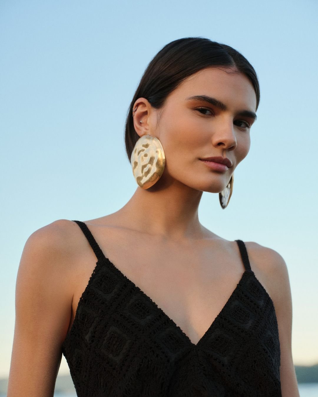 Mulher com brincos grandes dourados e vestido preto rendado, refletindo as tendências primavera verão.