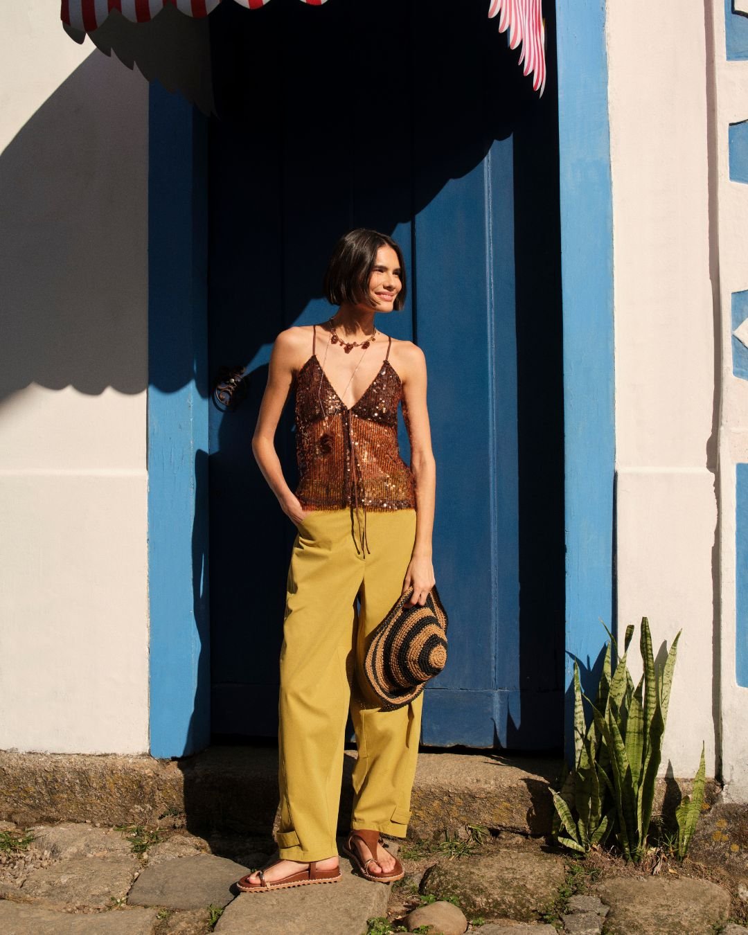 Pessoa em frente a uma porta azul, usando regata brilhante e calça amarela, segurando chapéu. Tendências de primavera verão.