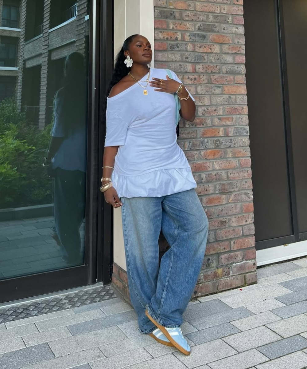 Pessoa de pé contra uma parede de tijolo, vestindo calça jeans e blusa branca. Tênis azul claro e bege.