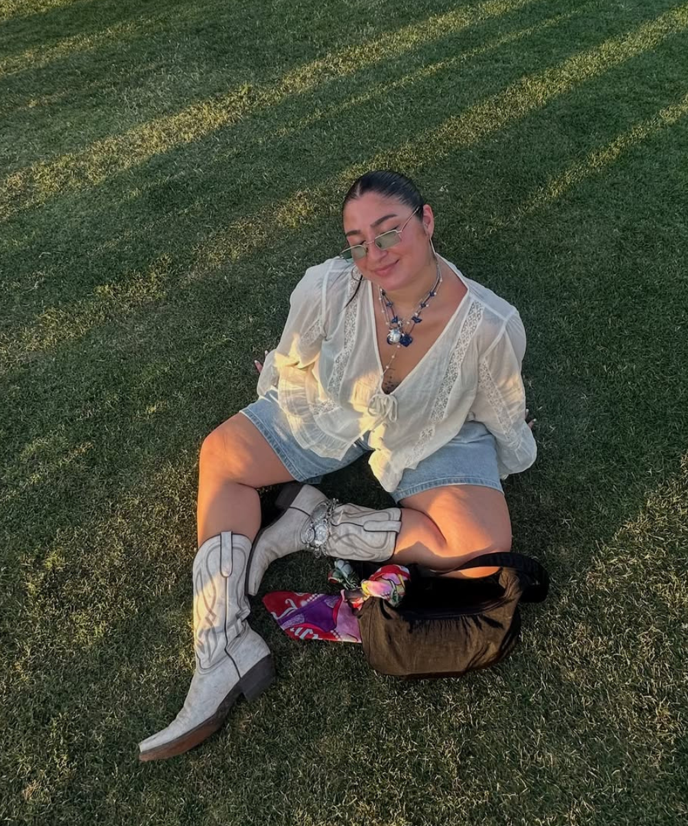 Pessoa sentada na grama usa blusa rendada, botas country e shorts. Look descontraído de festival.