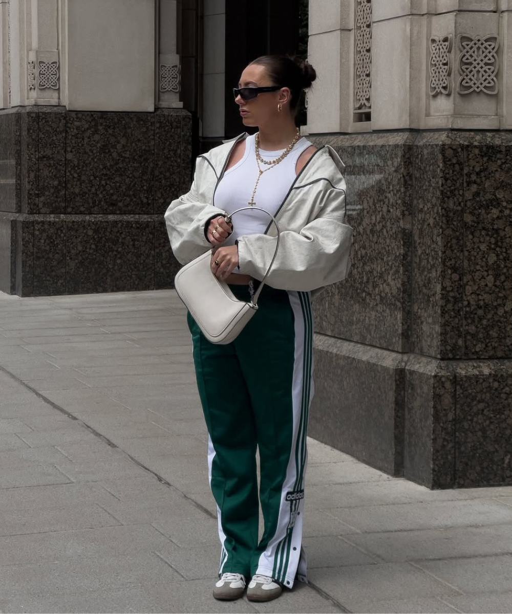 Mulher com jaqueta bege, regata branca, calça verde com listras, bolsa e óculos escuros.