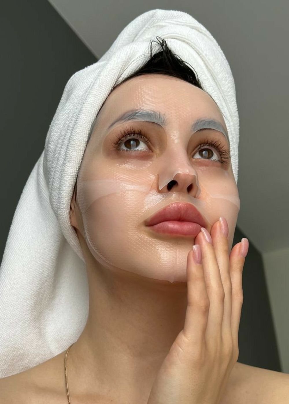 Pessoa com toalha na cabeça e máscara facial hidratante, olhando para cima, toca o rosto com a mão.