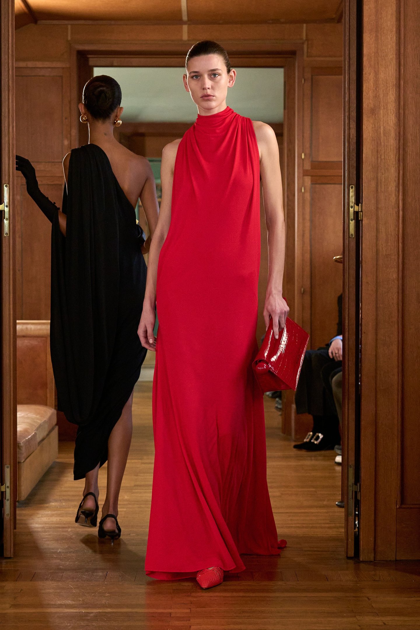 Modelo veste longo vermelho tomate e carrega bolsa combinando, enquanto outra em vestido preto ao fundo.
