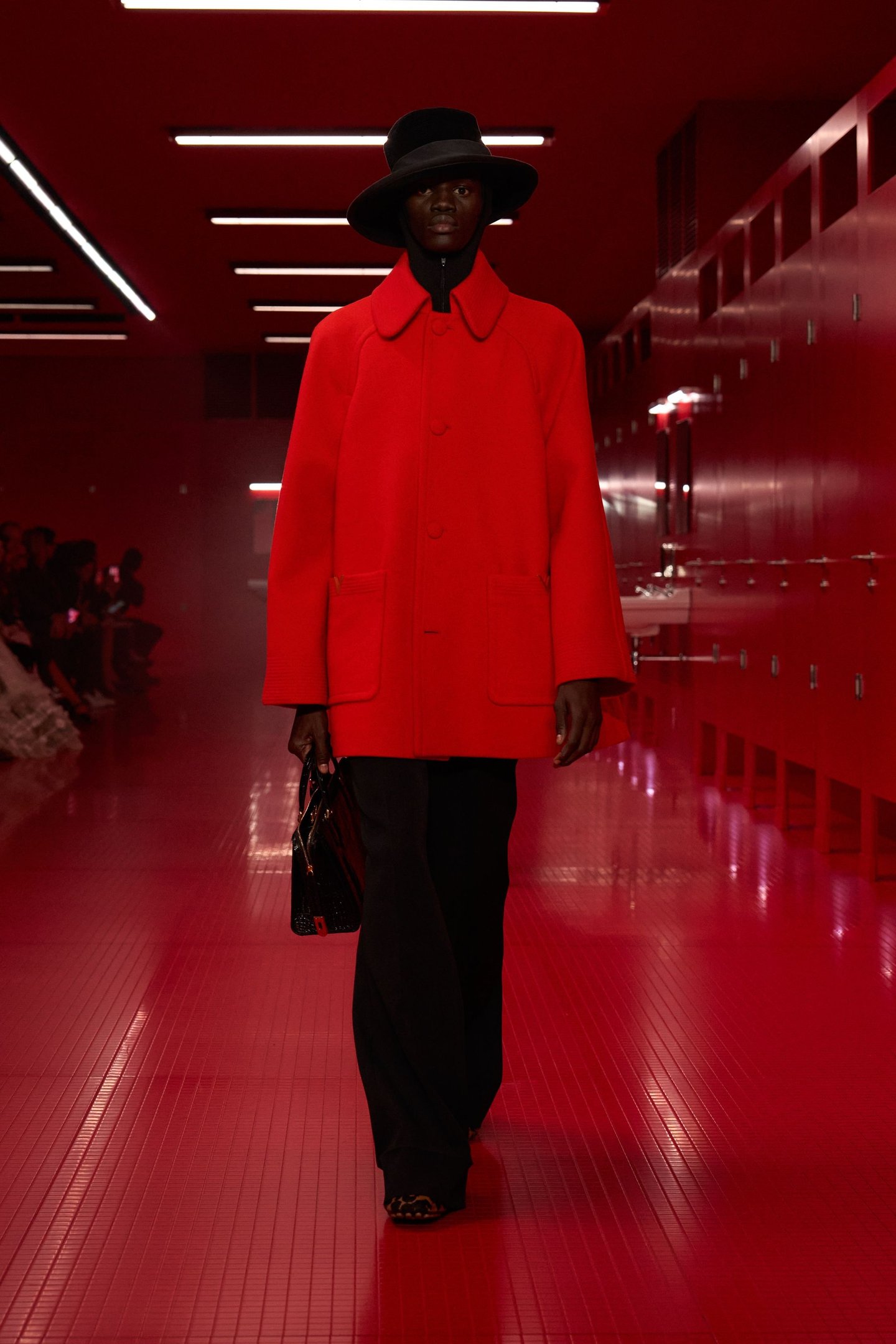 Modelo veste casaco vermelho tomate com chapéu preto em passarela vermelha iluminada.