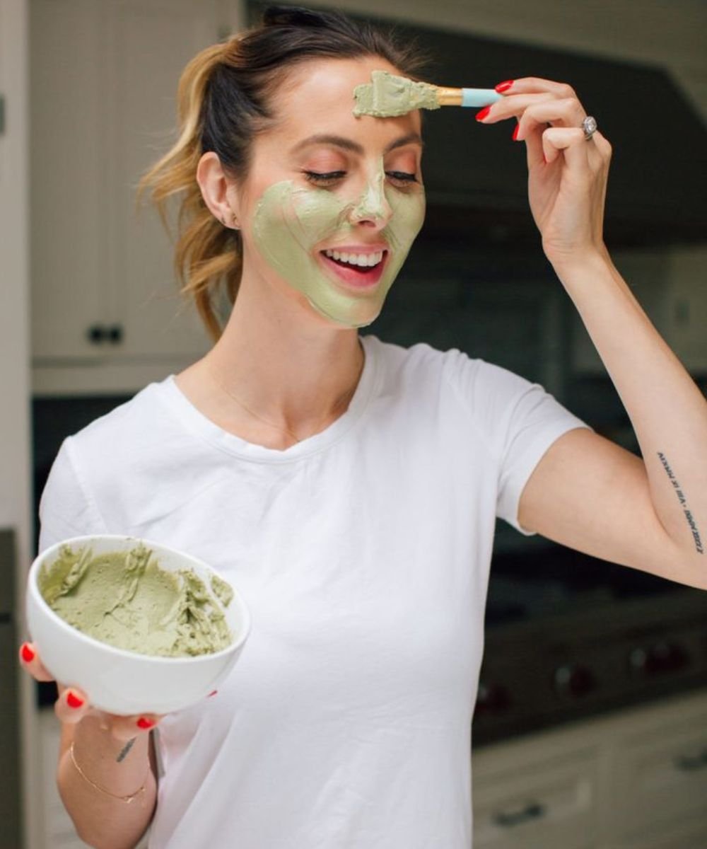 Mulher aplicando máscara facial verde para cuidados com a pele oleosa, segurando tigela e pincel.