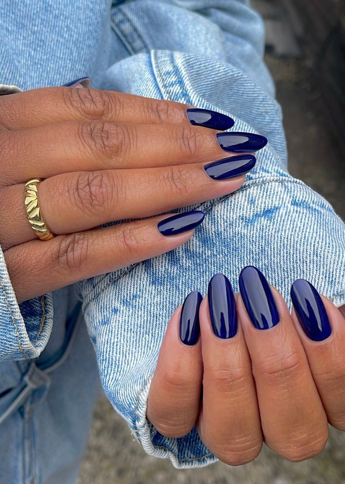 Unhas azul marinho, formato amendoado elegante, com anel dourado, em mão sobre jeans.