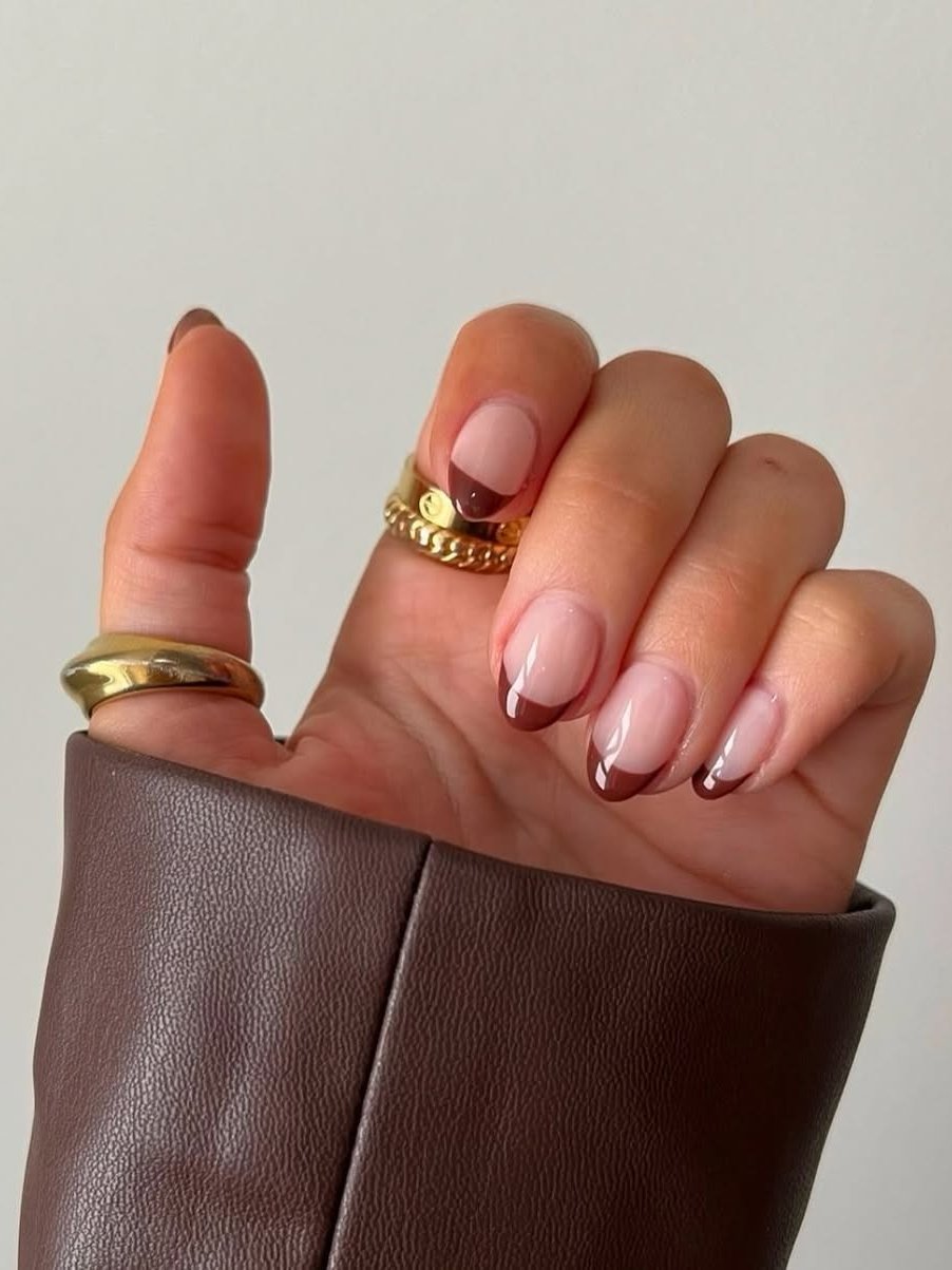 Unha amendoada elegante com bordas marrons, combinadas com anéis dourados e jaqueta marrom.
