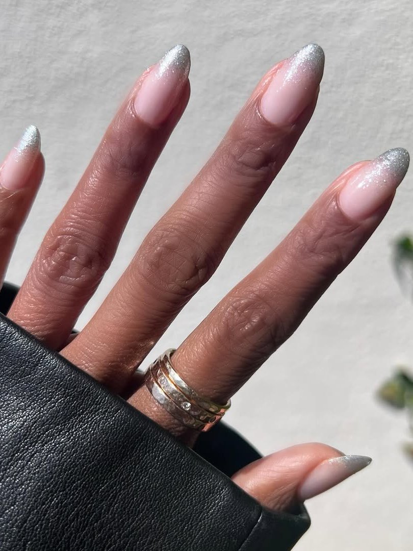 Unhas amendoada elegante com esmalte rosa e pontas prateadas, usando anéis dourados.