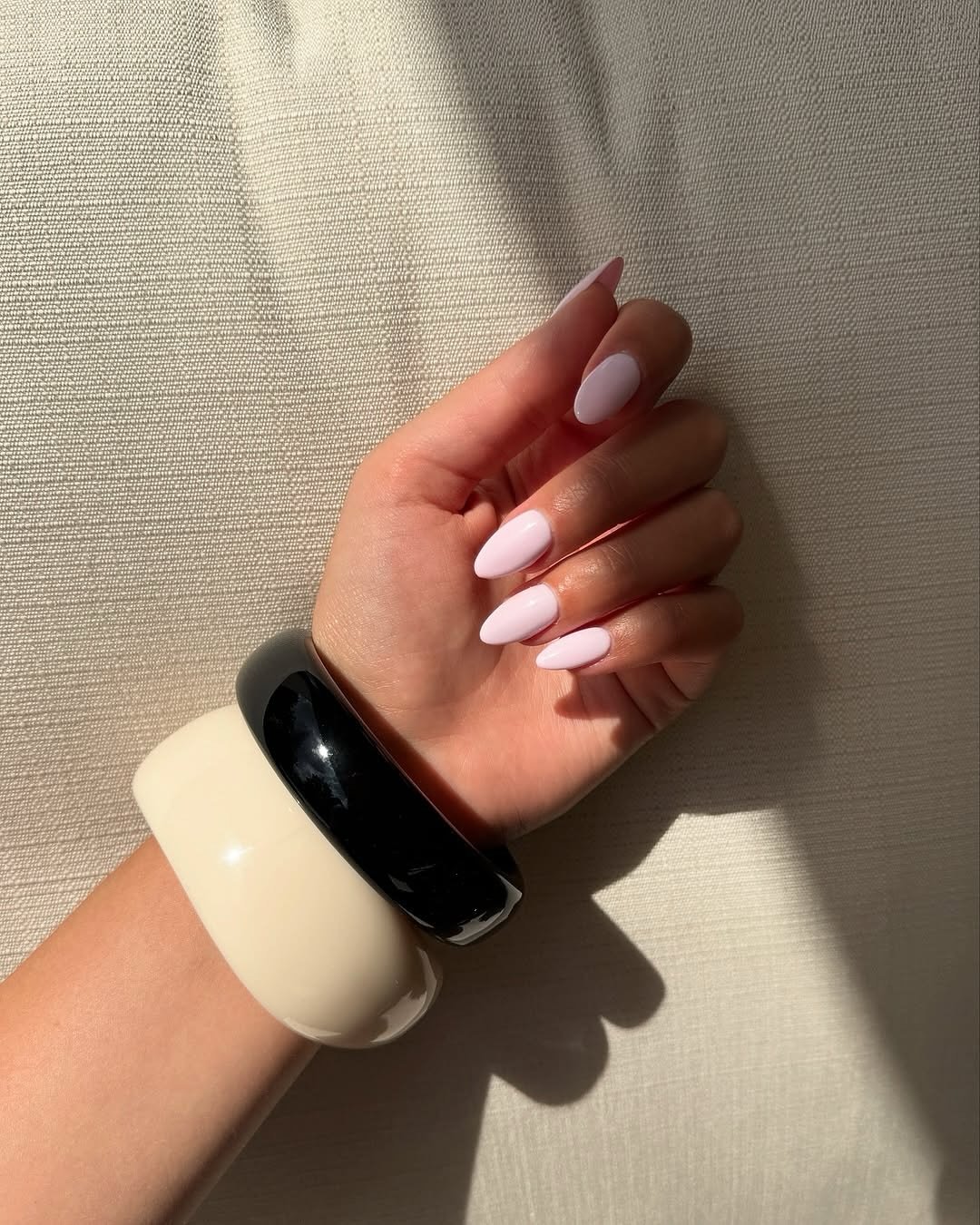 Unhas amendoada elegantes com esmalte claro, adornadas por pulseiras largas em tons de preto e creme.